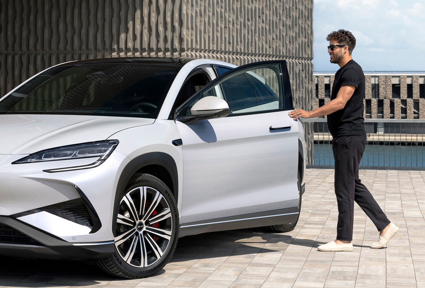 En mand med solbriller åbner døren i førersiden af en sølvfarvet SUV, der er parkeret på et asfalteret område i nærheden af en moderne bygning.
