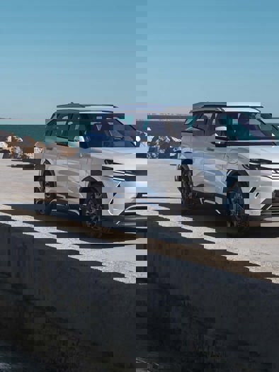 En hvid SUV er parkeret nær en klippekyst med havet i baggrunden under en klar himmel, hvilket skaber en naturskøn og eventyrlig atmosfære.