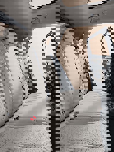 To beige bilsæder med tekstureret polstring, set fra bagsædet, vendt mod forruden hvor sollys strømmer ind og skaber en varm atmosfære.