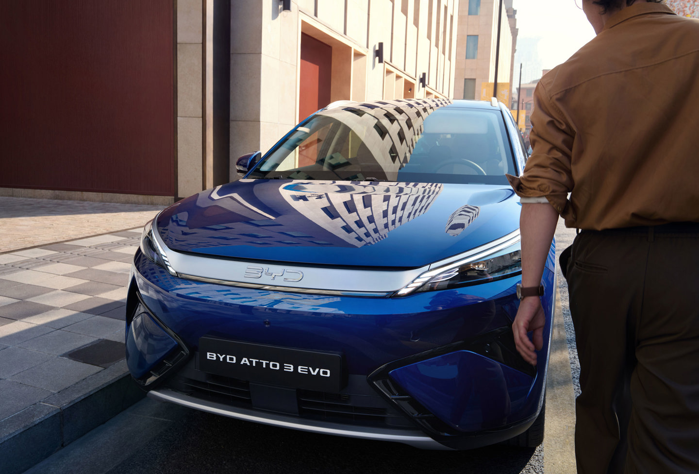 En person står ved siden af en blå BYD ATTO 3 EVO elbil, der er parkeret på en gade i byen i dagslys.