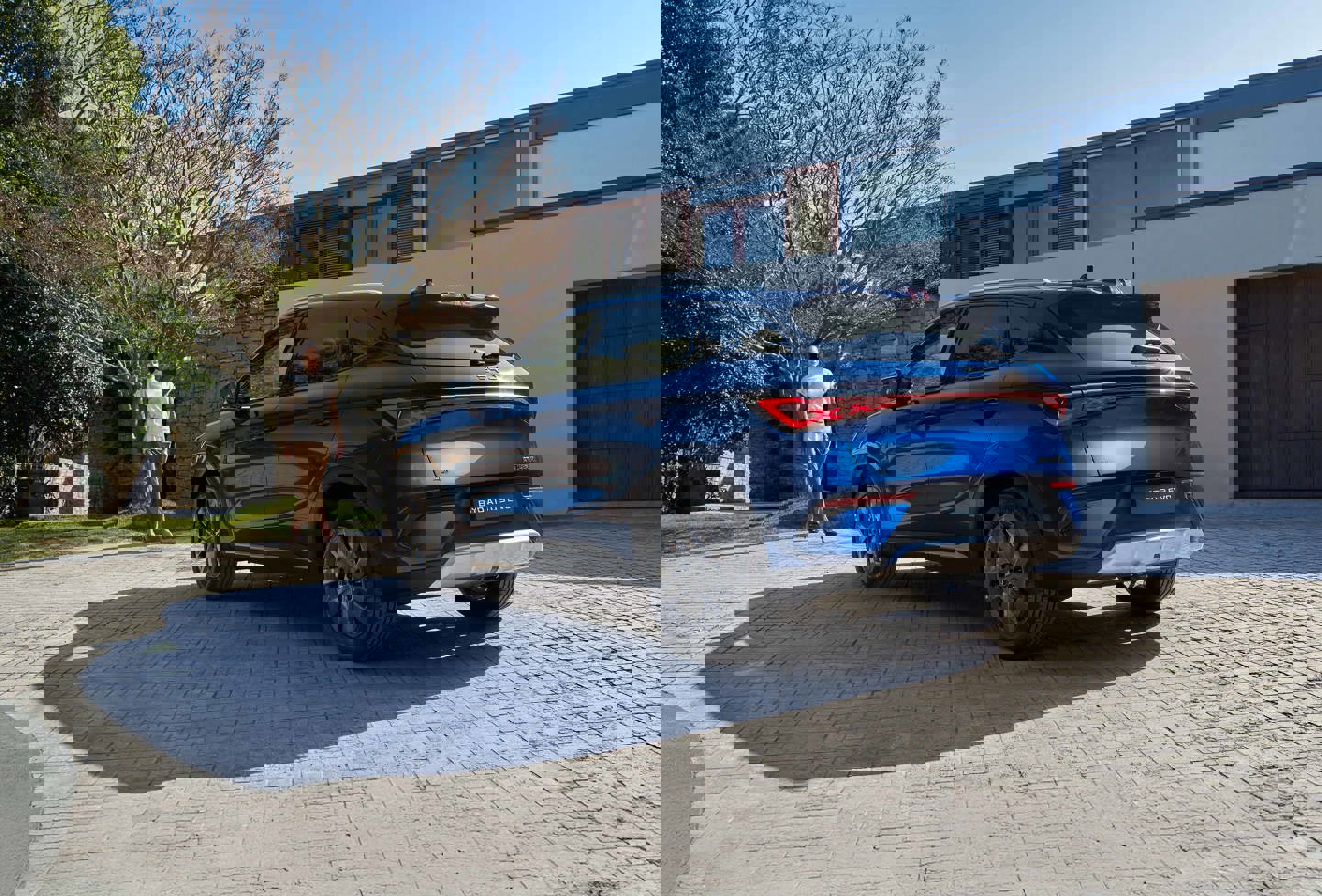 En blå SUV er parkeret på en stenbelagt indkørsel omgivet af bygninger og træer under en klar, solrig himmel, hvilket skaber en behagelig og indbydende scene.