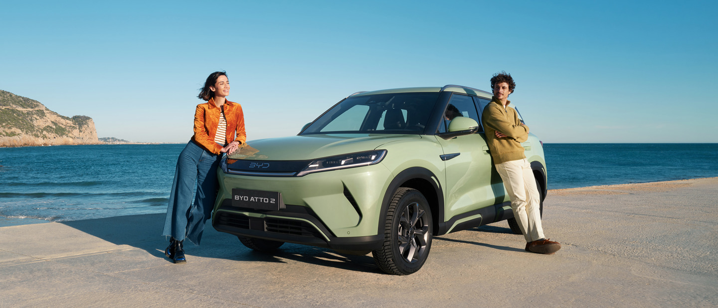 En kvinde og en mand læner sig op ad en lysegrøn BYD ATTO 2 SUV, der er parkeret ved havet under en klar blå himmel.