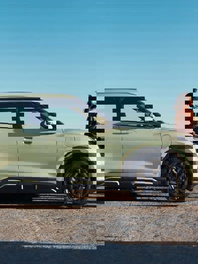 To personer står ved siden af en lysegrøn bil, der er parkeret nær et naturskønt område under en klar, blå himmel.